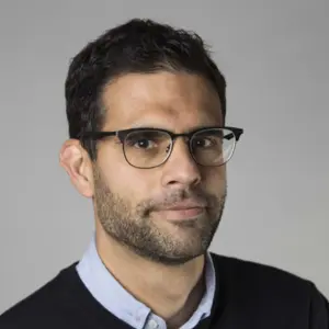 A man with short dark hair, a trimmed beard and black-rimmed glasses looks at the camera with a slight smile. He wears a black V-neck jumper over a light blue collared shirt. Plain grey background.